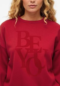 Sweatshirt rouge avec un grand imprimé en texte "SOIS TOI" dans différentes tailles et styles de police. Texture lisse, col rond, coupe décontractée.