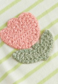 Fleur en crochet rose et feuille verte sur un tissu blanc avec des rayures vert clair. La broderie présente un motif texturé et tissé.