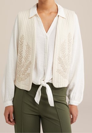 Vrouw draagt een witte blouse met textuur, gestrikt in de taille, een crèmekleurig doorgestikt vest met bladborduurwerk en olijfgroene broek.