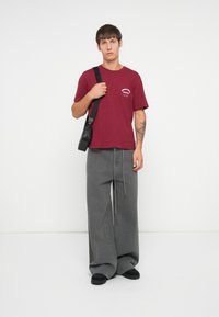 Bordeaux bomull t-skjorte med logo detalj, kombinert med løse grå jeans. Modellen bærer en svart skulderveske og har på seg svarte sko.