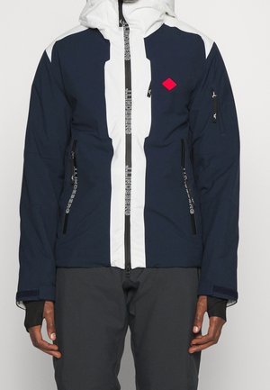 Granatowo-biała kurtka z przednim zamkiem błyskawicznym, dwoma zamykanymi kieszeniami, otworami na kciuki w mankietach oraz czerwonym logo w kształcie diamentu na piersi.