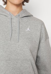 Personne portant un sweat à capuche gris avec un logo Jumpman blanc sur la poitrine et des cordons pendants.