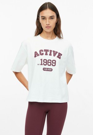 Jonge vrouw draagt een wit oversized T-shirt met de tekst "ACTIVE 1969 S.OLIVER" en bordeauxrode leggings, staand tegen een effen achtergrond.