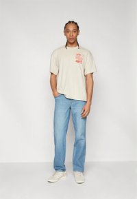 Μπεζ βαμβακερό μπλουζάκι με κόκκινο γραφικό στο αριστερό στήθος, σετ με ανοιχτό μπλε τζιν παντελόνι και λευκά αθλητικά παπούτσια. Απλός, καθημερινός σχεδιασμός.