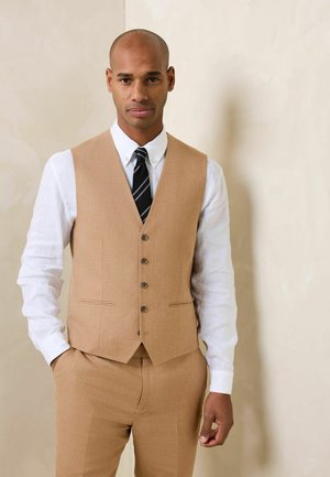 Mann trägt eine hellbraune Anzugweste und Hose mit weißem Hemd und schwarz gestreifter Krawatte, steht vor einem beigen Hintergrund.