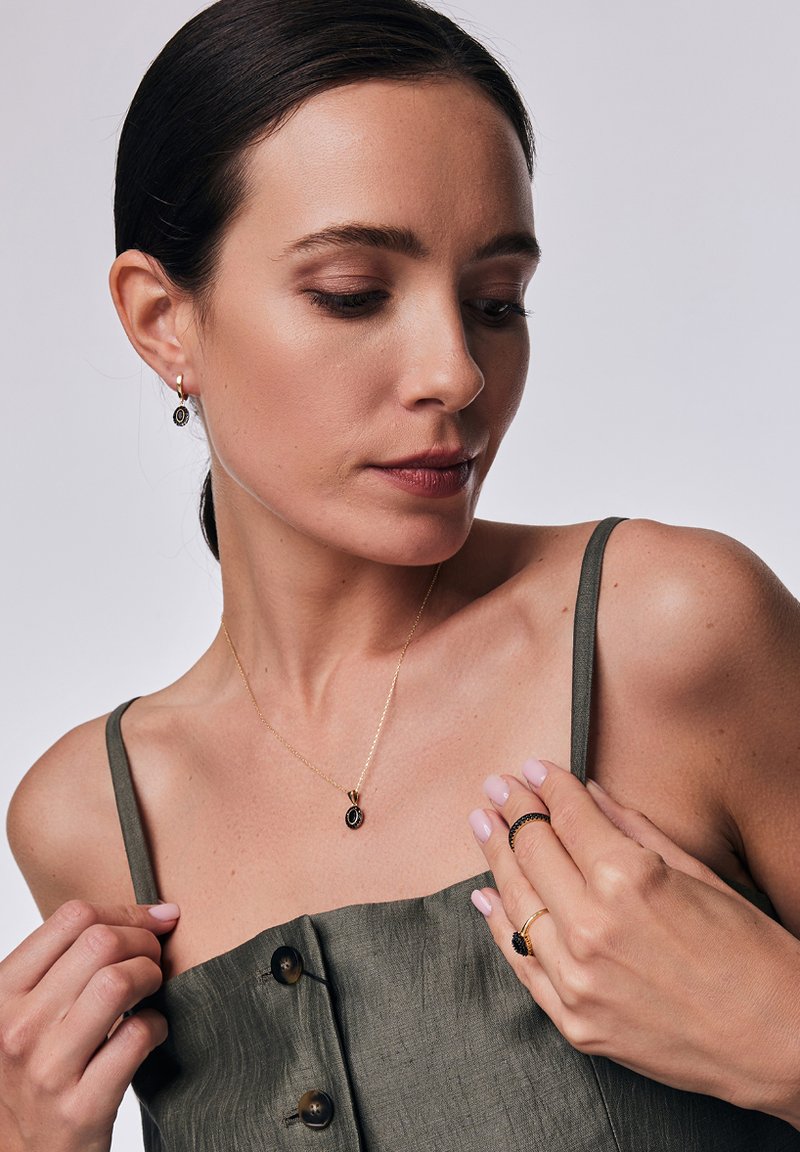 Złoty naszyjnik z czarnym wisiorkiem, małe złote kolczyki w kształcie kółek oraz pierścionki w warstwie. Modelka nosi zielony strój z guzikami. Miękkie oświetlenie.