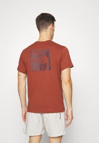 Camiseta de algodón color óxido con un estampado gráfico en la espalda, cuello redondeado y mangas cortas, combinada con pantalones cortos claros.