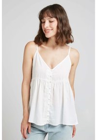 Camisole en lin blanc avec de fines bretelles, décolleté en V, détail de boutons sur le devant et un bas arrondi ample et évasé. Assorti avec un jean clair.