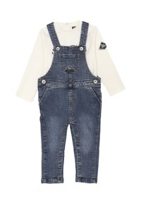 Salopette in denim con tasca frontale, spalline regolabili e bottoni argentati. Abbinata a una maglietta a maniche lunghe bianca con un'etichetta logo nera.