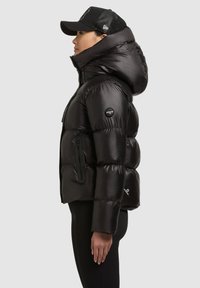 Musta toppatakki, jossa korkea kaulus, kiiltävä pinta, sivuvetoketjut ja hihassa logopläntti. Yhdistetty mustaan lippalakkiin.