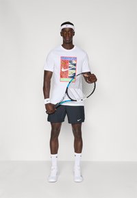 Tricou grafic alb cu modele multicolore, pantaloni şort negri, adidaşi albi şi o rachetă. Accessorizat cu o bandă albă pentru cap.