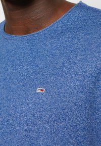 Camiseta azul de manga corta con una tela texturizada. Presenta un pequeño logotipo bordado en rojo, blanco y azul marino en la parte izquierda del pecho.