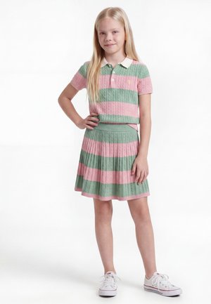 Young girl standing with hand on hip, wearing pink and green striped short-sleeve top and matching skirt, white sneakers, on a white background.
