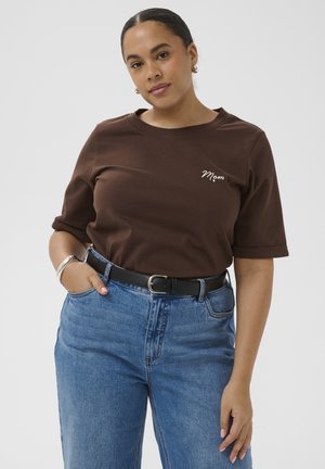 Kobieta z ciemnymi włosami w brązowej koszulce z napisem "Mama", niebieskich, wysokich dżinsach, czarnym pasku i srebrnej bransoletce, pozuje na tle jednolitego jasnego tła.