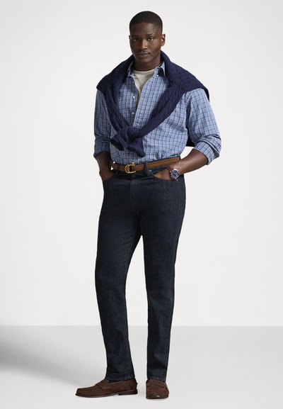 Modra srajca s karo vzorcem, pod njo siva majica, mornarsko modra jopica okoli ramen, temne jeans hlače, rjavi mokasini in ročna ura.