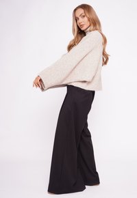 Kobieta z długimi falowanymi włosami, ubrana w beżowy, oversize'owy sweter oraz szerokie czarne spodnie, stoi bokiem na jednolitym tle.