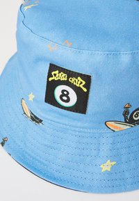 Kék vászon sapka, nyolcas billiárdgolyóval díszítve, furcsa grafikai elemekkel nyaralókról, valamint sárga és narancssárga csillagokkal.