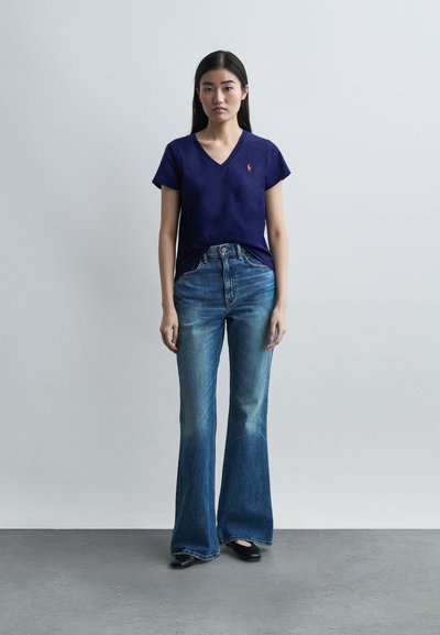 Νεαρή γυναίκα που στέκεται μπροστά σε έναν απλό τοίχο, φορώντας μπλε μπλούζα με V-λαιμό, καμπύλα μπλε τζιν και μαύρα παπούτσια, κοιτάζοντας μπροστά με ουδέτερη έκφραση.