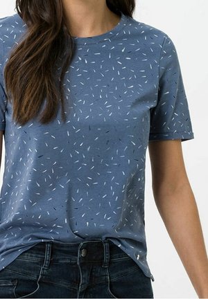 Frau trägt ein blaues Kurzarmshirt mit kleinen weißen und schwarzen Strichmustern und dunkelblaue Jeans.