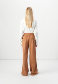 Loosely fitting brown pants with an elastic waistband, made of soft fabric, paired with a white long-sleeve shirt. Rear view shown.