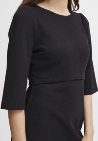 Svart texturerad klänning med trekvartslånga ärmar, med en åtsittande bodice och en horisontell söm över bröstet. Slät, vävd material.