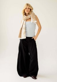 Beige en crèmekleurige geprinte sjaal hangt over een lichtblauw tanktop, gecombineerd met losse zwarte broeken en donkerbruine schoenen.