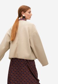 Mujer con cabello largo y rojo en una coleta baja, lleva pendientes grandes de oro y negro, una chaqueta beige de gran tamaño y un vestido con un estampado.