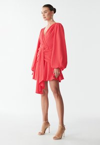 Rotes, strukturiertes Wickelkleid mit langen Ballonärmeln, einem rüschenbesetzten Saum und einem Bindeband in der Taille. Kombiniert mit goldenen Riemchen-High Heels.