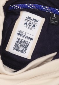 Étiquette d'un vêtement, présentant un tissu crème avec des accents bleu marine foncé, une bordure tressée bleue, et un texte imprimé incluant un code QR.