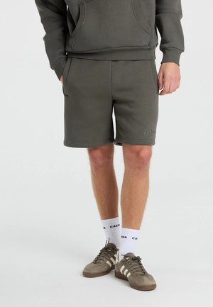 Mand iført mørkegrønne casual shorts, matchende hættetrøje, hvide sokker med tekst og brune sneakers med hvide striber.