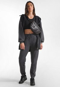 Schwarzer Hoodie mit oversized Schnitt, kombiniert mit passenden Jogginghosen und schwarzen Sneakern. Eine Umhängetasche setzt einen glänzenden, strukturierten Akzent.
