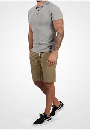 Mann trägt graues kurzärmeliges Poloshirt, beige Shorts mit geflochtenem Gürtel, schwarz-weiße Sneaker, steht mit entspannten Händen, Tätowierung am rechten Unterarm sichtbar.