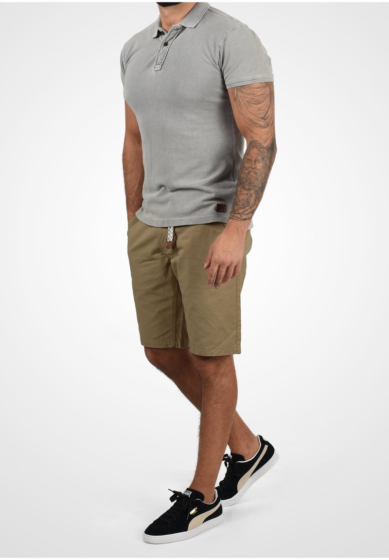 Homme portant un polo gris à manches courtes, un short beige avec une ceinture tressée, des baskets noires et blanches, debout les mains détendues, tatouage visible sur l'avant-bras droit.