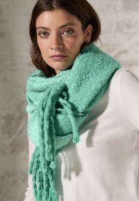 Sciarpa turchese realizzata in morbido tessuto testurizzato. Presenta dettagli frangiati e un design drappeggiato, indossata sopra una maglietta bianca su uno sfondo neutro.
