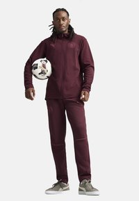 Tuta color granata con giacca con zip e pantaloni, caratterizzata da un logo, cuciture di contrasto e tessuto liscio. La persona tiene un pallone da calcio.