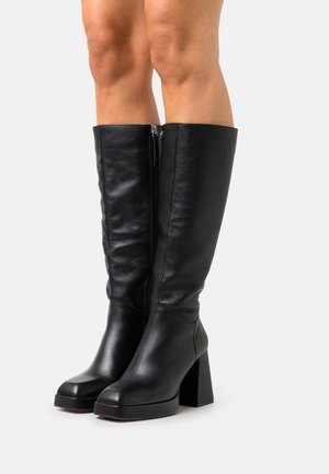Černé kožené boty ke kolenům s masivními podpatky, nošené na holých nohou, zobrazené na jednotném světle pozadí.