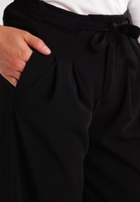 Pantalon noir plissé en tissu texturé, avec une taille à cordon de serrage et des poches latérales pour un usage pratique.