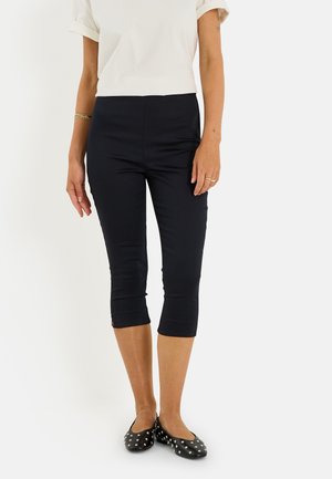 Femme portant un pantalon capri noir, un haut blanc à manches courtes, des ballerines noires avec des clous argentés, debout devant un fond blanc uni.