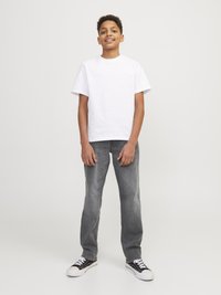 White cotton t-shirt, gray denim jeans, and black sneakers with white accents. Model stands upright, smiling on a plain background.