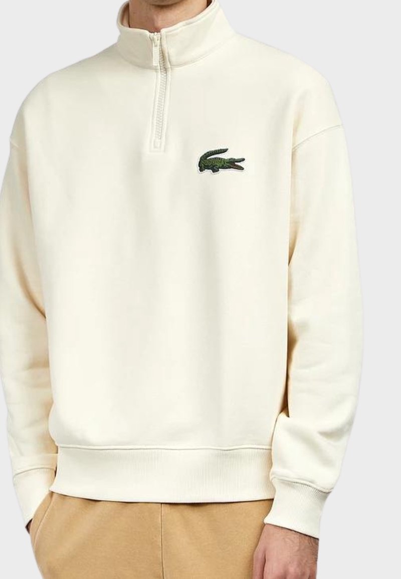 Kremowy sweatshirt z zamkiem na szyi, z ribbami na dolnym hemie i mankietach. Posiada zielone haftowane logo krokodyla na lewej piersi. Miękka faktura.