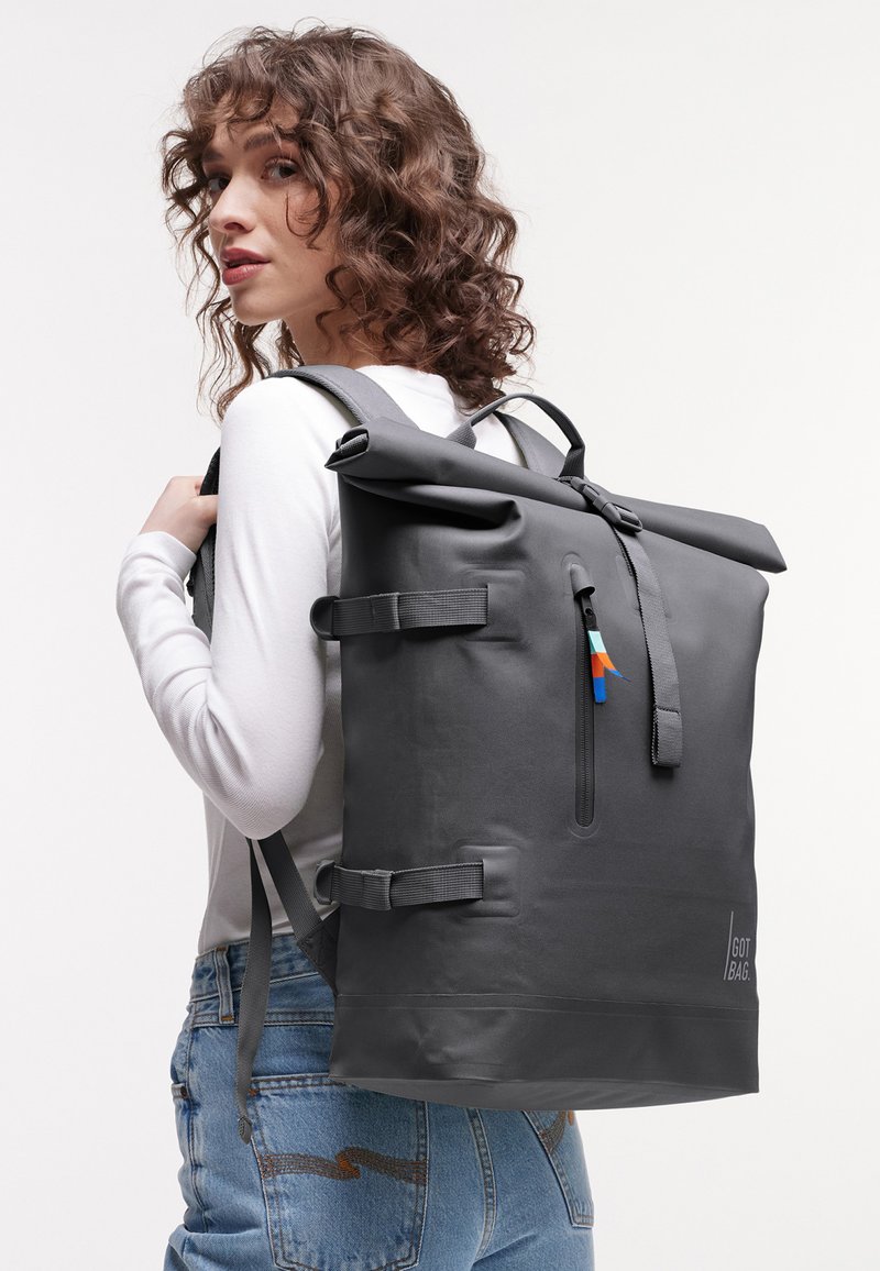 Giovane donna con capelli ricci che indossa una maglietta bianca e jeans, porta uno zaino nero grande con chiusura a rotolo e cinghie laterali.