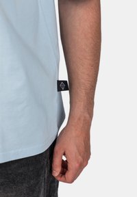 Světle modré bavlněné tričko s krátkými rukávy a černou cedulkou se diamantovým logem, umístěné vedle ruky odpočívající na tmavých džínech.