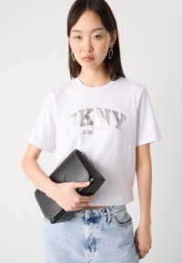 Balta medvilnės marškinėliai su sidabriniu "DKNY NEW YORK" spaudiniu, derinami su juoda odine rankine, pasižyminčia balta siuvinėta apdaila ir stačiakampiu siluetu.