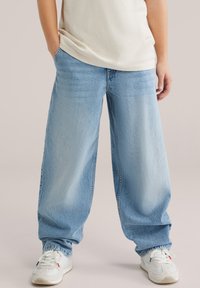 Helehallid teksapüksid laia säärega ja lõdva istuvusega, millel on traditsiooniline viie taskuga disain ja õrn tuhmumine. Kandmisel valgete tossudega.