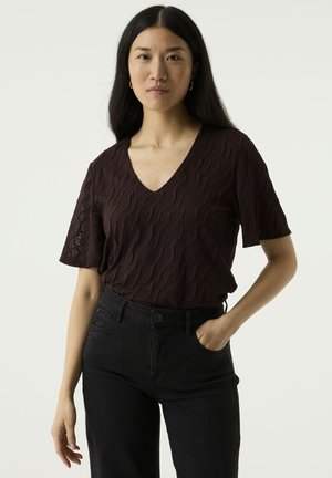 Femme aux longs cheveux noirs portant un haut texturé sombre à col en V et manches courtes ainsi qu'un pantalon noir, une main dans la poche, expression neutre.