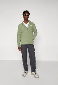 Vihreä fleecetakki vetoketjulla, harmaat housut ja vaaleanharmaat lenkkarit. Huomattavat valkoiset logoväritykset rinnassa ja reidessä, jotka täydentävät aurinkolasit.