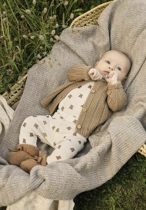 Pleteni bež cardigan preko kreme outfita s printom medvjedića, meke smeđe čizmice, smještene u teksturiranoj sivoj deki na travi.