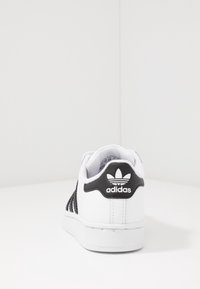 Basket blanche avec des accents noirs et des côtés en mesh. Dispose d'une pointe arrondie, d'un col rembourré et d'un logo Adidas noir distinct au niveau du talon.