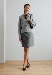 Cardigan gris à côtes avec des boutons, associé à une jupe rayée grise. Le modèle tient un sac à main noir et porte des chaussures plates noires ainsi qu'un foulard.