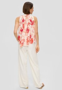 Ärmellose, leichte Bluse in Pastellrosa mit rotem Blumenmuster; bietet ein plissiertes Design und Bindebänder am Rücken. Kombiniert mit weißen, weit geschnittenen Hosen.
